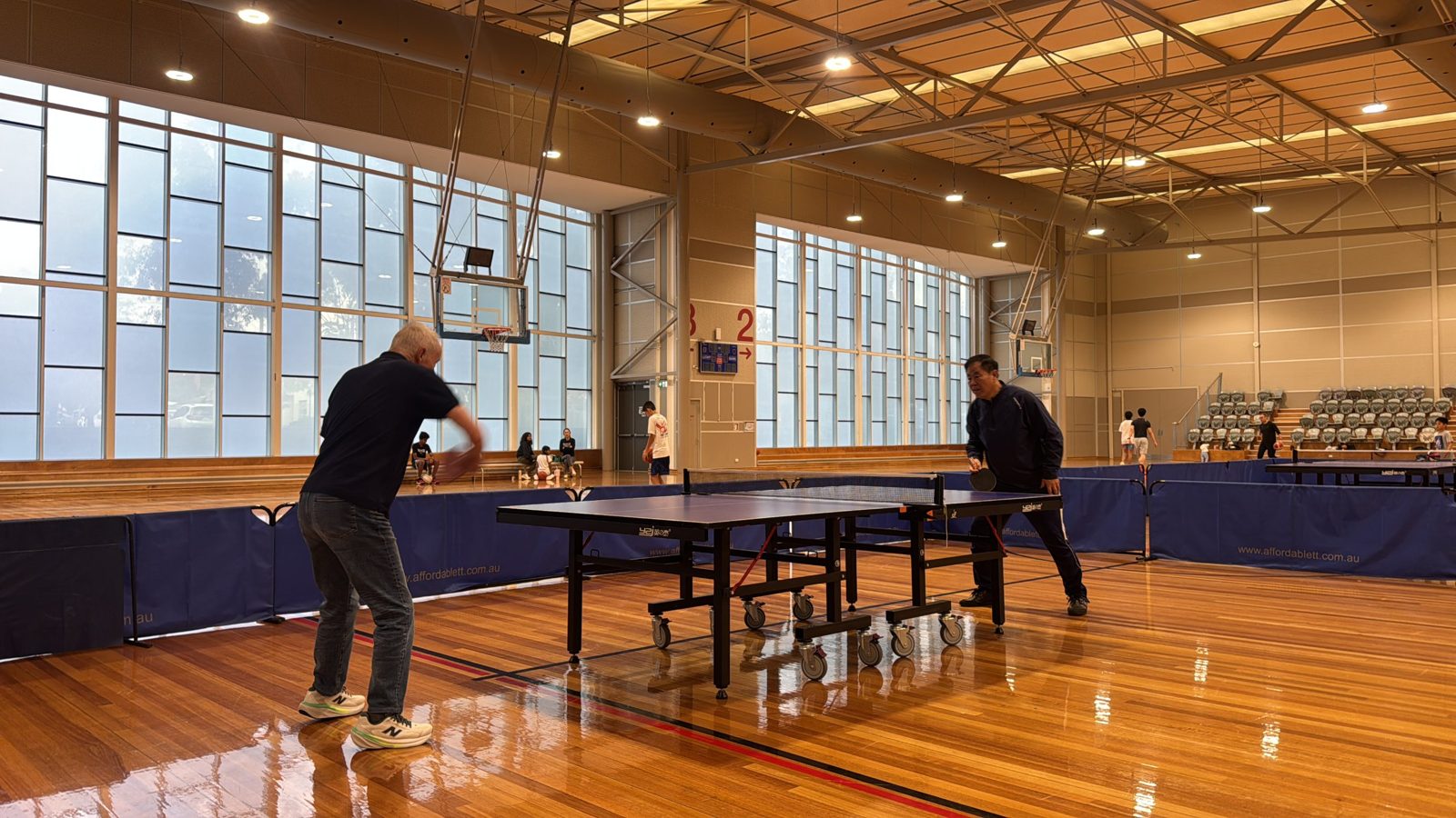 Two members playing singles on a blue table under the stadium windows